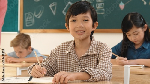 Asian smart boy smiling to camera while student writing answer in answer sheet. Multicultural student doing classwork or test while caucasian teacher checking student homework at classroom. Pedagogy.