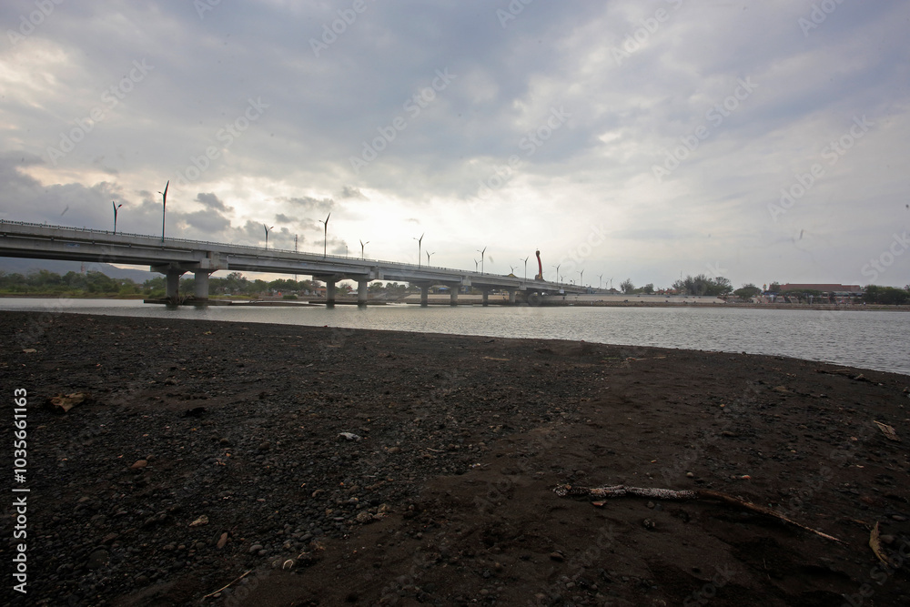 The image shows a large concrete bridge spanning over a river under a cloudy sky, with calm water beneath. Streetlights line the bridge, creating an urban atmosphere around the quiet riverside area The image shows a large concrete bridge spanning over a river under a cloudy sky, with calm water beneath. Streetlights line the bridge, creating an urban atmosphere around the quiet riverside area