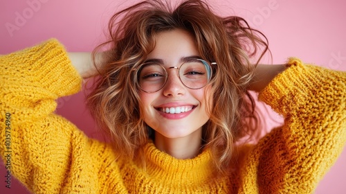 A person with a radiant smile and stylish glasses accessorizes a cozy yellow sweater, standing against a pink background and radiating happiness. photo