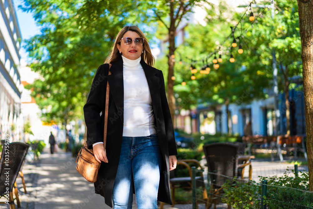 Middle aged elegant woman in black coat, jeans, cream turtleneck, leather handbag and sunglasses walking on sidewalk in city in autumn. Everyday life in city. Front view. Middle aged elegant woman in black coat, jeans, cream turtleneck, leather handbag and sunglasses walking on sidewalk in city in autumn. Everyday life in city. Front view.
