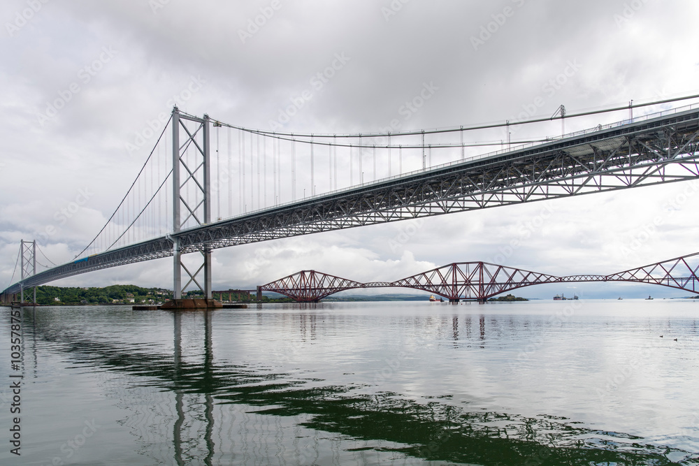 Fototapeta premium Ship sailing under the red Forth cantilever railway bridge across the Firth of Forth, Queensferry, UK and new Forth Road Bridge crossing in top of image near Edinburgh