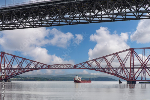 Ship sailing under the red Forth cantilever railway bridge across the Firth of Forth, Queensferry, UK and new Forth Road Bridge crossing in top of image near Edinburgh
