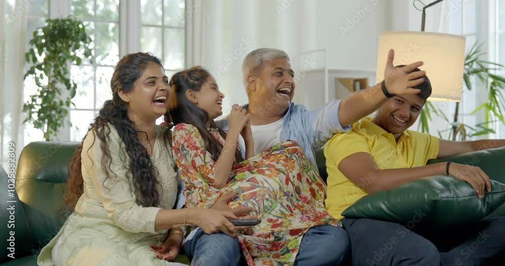 Indian Asian family of four, parents in their forties and two kids, sitting closely on a sofa, laughing out loud while watching television together, enjoying a fun-filled family evening at home Indian Asian family of four, parents in their forties and two kids, sitting closely on a sofa, laughing out loud while watching television together, enjoying a fun-filled family evening at home