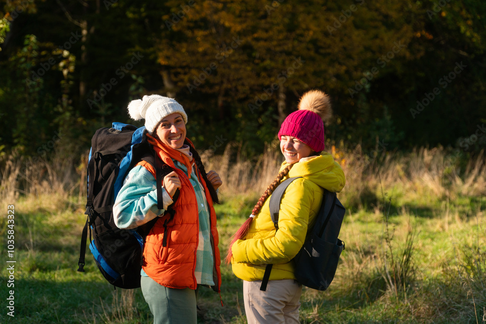  family walking in autumn forest with backpacks, boy in yellow jacket, girl in pink jacket, mother in orange sleeveless jacket, family in hat with pompoms, gen z