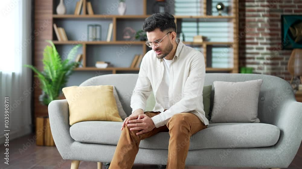 Handsome young adult man suffers from painful severe knee joint pain while sitting on sofa in living room at home. Upset bearded male massages leg muscles, could not walk, illness, cramps, rheumatism Handsome young adult man suffers from painful severe knee joint pain while sitting on sofa in living room at home. Upset bearded male massages leg muscles, could not walk, illness, cramps, rheumatism