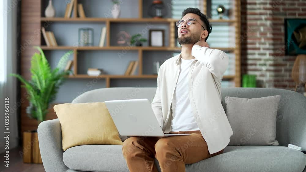 Tired young adult bearded man suffering from neck pain while working on laptop sitting on sofa in living room at home. Upset male in glasses feels discomfort, massages and rubs sore muscles, stretches Tired young adult bearded man suffering from neck pain while working on laptop sitting on sofa in living room at home. Upset male in glasses feels discomfort, massages and rubs sore muscles, stretches