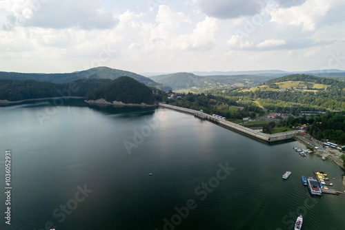 Aerial view of a tranquil lake surrounded by lush green hills and a dam structure, showcasing serene natural beauty