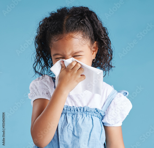 Girl, child and sneeze with tissue in studio, sick or cold with mucus, cleaning nose and hygiene by blue background. Girl, allergies or hay fever with paper towel for infection, virus and bacteria