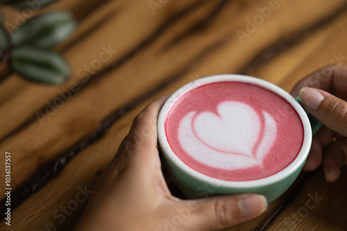 Young woman's hand holds a cup of matcha tea with raspberry or red berries, heart-shaped milk art, cappuccino coffee with red foam and heart, wooden table and decoration with plants, vegan hot drink