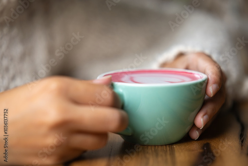 Young woman's hand holds a cup of matcha tea with raspberry or red berries, heart-shaped milk art, cappuccino coffee with red foam and heart, wooden table and decoration with plants, vegan hot drink