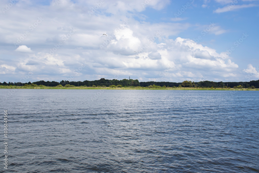 A serene view of calm waters reflecting the sky with scattered clouds and lush greenery in the background. A serene view of calm waters reflecting the sky with scattered clouds and lush greenery in the background.
