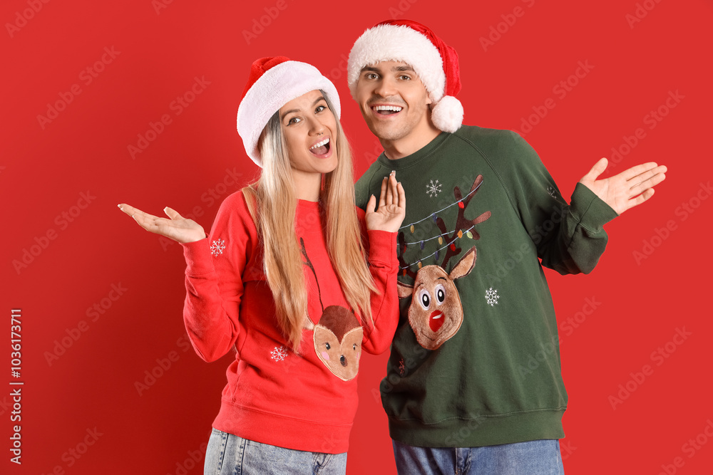 Young couple in Santa hats and Christmas sweaters on red background Young couple in Santa hats and Christmas sweaters on red background