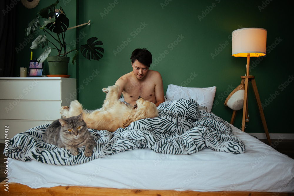 A young man enjoys a tranquil morning in bed, petting a golden dog while a soft gray cat lounges nearby. A perfect snapshot of relaxation and the bond between pets and their owner.