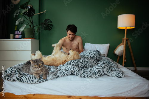 Obraz na płótnie A young man enjoys a tranquil morning in bed, petting a golden dog while a soft gray cat lounges nearby. A perfect snapshot of relaxation and the bond between pets and their owner. w salonie