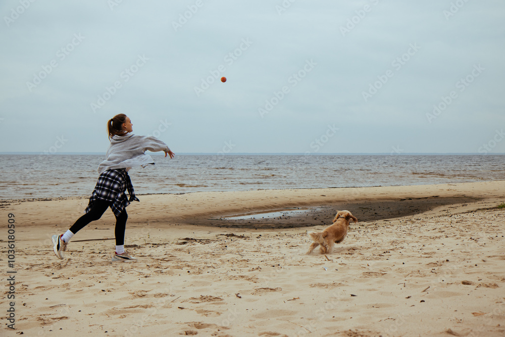 Young woman playing fetch with her dog on the beach. The dog eagerly chases a ball, creating a joyful and energetic scene by the sea. Casual autumn attire and playful outdoor activity.