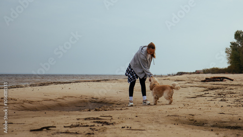 Obraz na płótnie Young woman enjoying a game of fetch with her dog by the sea. The dog runs after a ball, while the girl smiles, reflecting joy and companionship in a beachside environment. w salonie