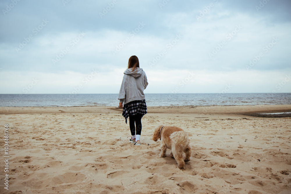 Young woman and her dog explore a quiet beach, both enjoying the fresh air and scenic ocean views. Casual autumn fashion and a tranquil moment in nature.