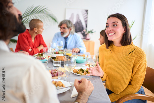 Young Caucasian beautiful woman smiling with cup of white wine in her hand sitting at table at family meal indoor. Multi-generational people gathered enjoying food in weekend festive event at home