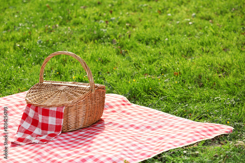 Fototapeta Naklejka Na Ścianę i Meble -  One picnic wicker basket with checkered napkin and blanket on green grass. Space for text