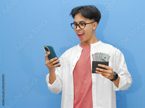 Portrait of a happy Asian man holding a wallet full of money and using a mobile phone with an amazed expression on an isolated blue background.