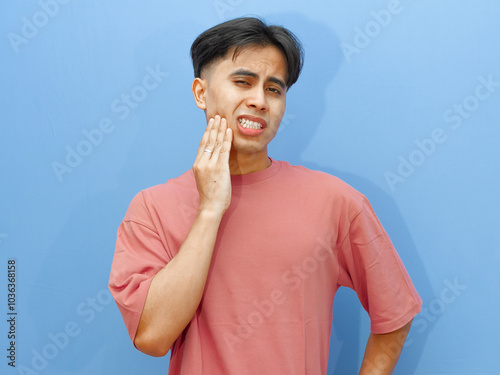 A young Asian man suffering from a painful toothache, holding his cheek with a sad expression, isolated on a blue background. Dental and oral care concept.