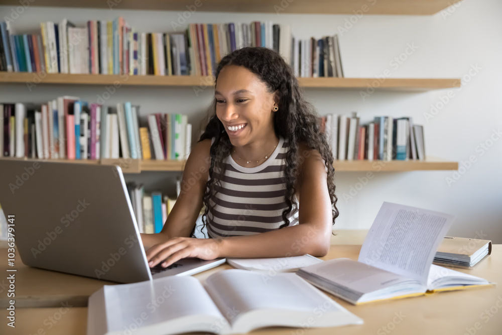 Cheerful young African American student girl studying in public university library, browsing Internet on laptop, working on research paper, typing, smiling, laughing, sitting at table with open books Cheerful young African American student girl studying in public university library, browsing Internet on laptop, working on research paper, typing, smiling, laughing, sitting at table with open books