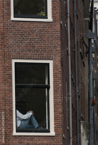 Person in white sweater and blue jeans sitting on window sill of a brick building on Brouwersgracht photo