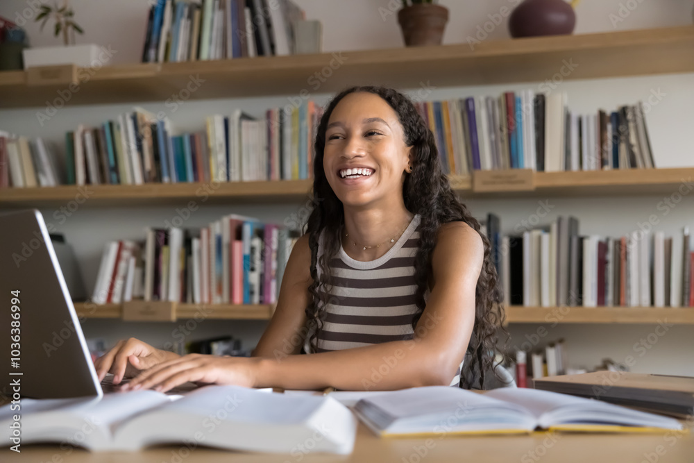 Happy fresh African student girl working hard in library, enjoying learning process, typing on laptop computer at table with heap of open books, looking away, smiling, laughing Happy fresh African student girl working hard in library, enjoying learning process, typing on laptop computer at table with heap of open books, looking away, smiling, laughing