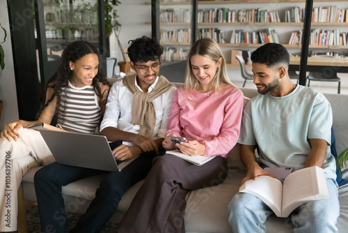 Multiethnic team of student friends discussing online learning content on gadgets, sitting close on couch, meeting in university library, educational environment, talking, smiling, laughing