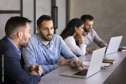 Two businessmen discussing professional online content at laptop, sitting at large co-working table, talking and working at computer, meeting for teamwork, collaboration, help