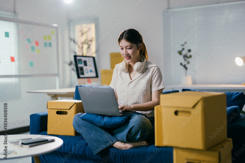 Young asian female entrepreneur is sitting on a blue couch and working on her laptop, surrounded by cardboard boxes, in her new, modern apartment Young asian female entrepreneur is sitting on a blue couch and working on her laptop, surrounded by cardboard boxes, in her new, modern apartment