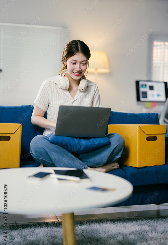Young asian woman is working from home, sitting on a blue sofa with her laptop on her lap. She is wearing headphones and smiling Young asian woman is working from home, sitting on a blue sofa with her laptop on her lap. She is wearing headphones and smiling
