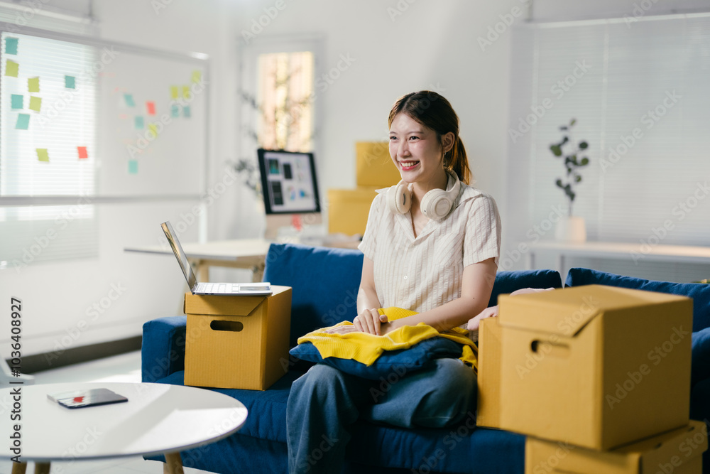 Excited asian woman unpacks boxes in bright new apartment, setting up home with laptop and headphones in sunny living room. Joyful moving day moment