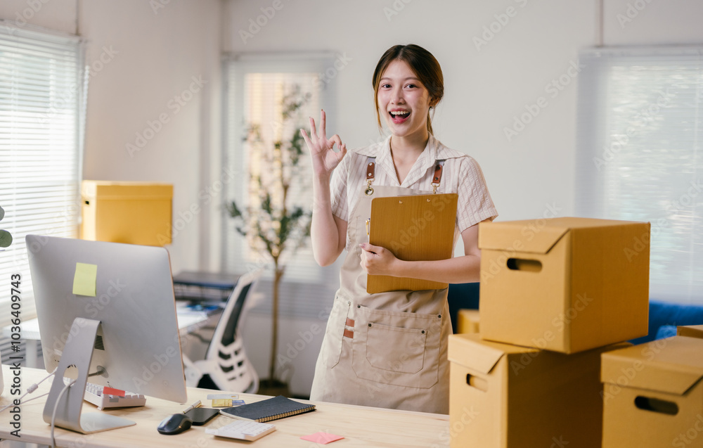 Young asian female entrepreneur is smiling and showing the ok sign while holding a clipboard in her home office, surrounded by cardboard boxes Young asian female entrepreneur is smiling and showing the ok sign while holding a clipboard in her home office, surrounded by cardboard boxes