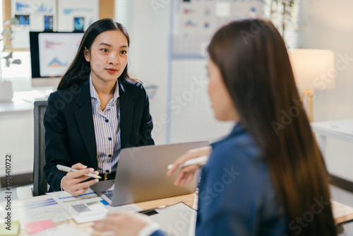 Two businesswomen having a discussion about work while sitting at a desk in an office. They are working together on a project