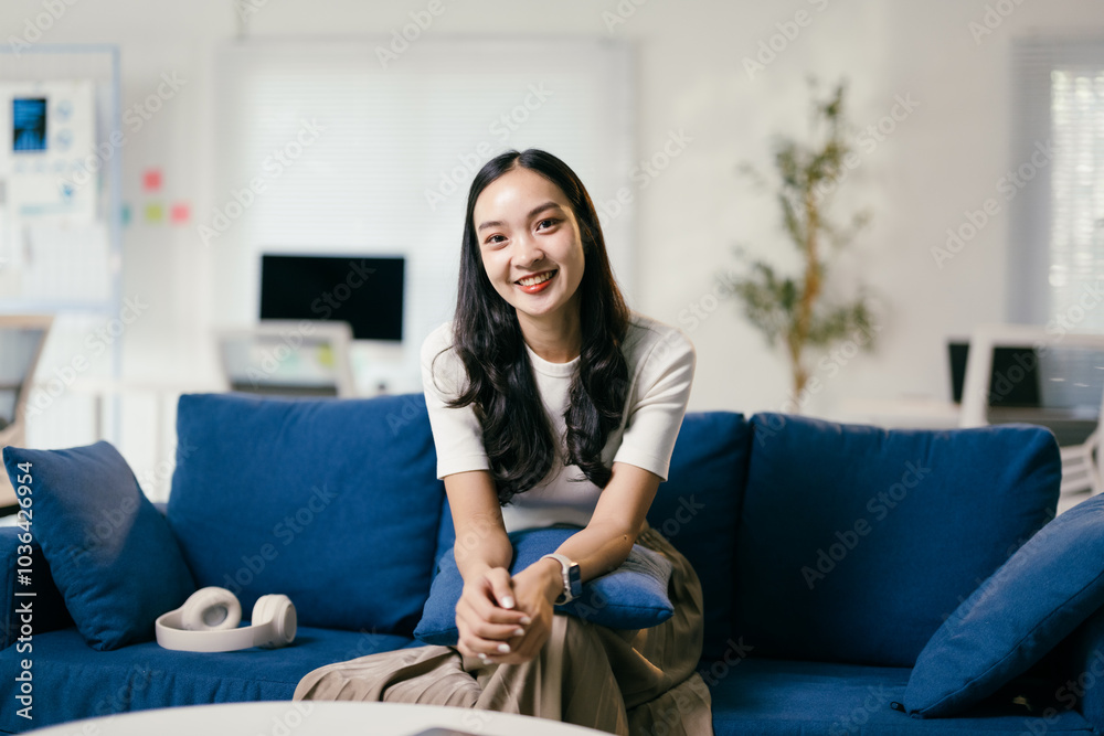 Young asian woman is sitting on a blue sofa in a modern office, smiling. She is wearing a white shirt and has long black hair Young asian woman is sitting on a blue sofa in a modern office, smiling. She is wearing a white shirt and has long black hair