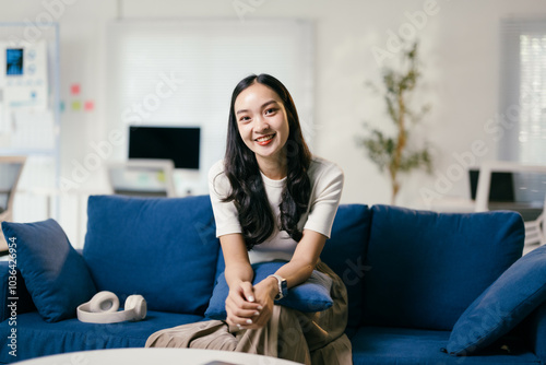 Young asian woman is sitting on a blue sofa in a modern office, smiling. She is wearing a white shirt and has long black hair