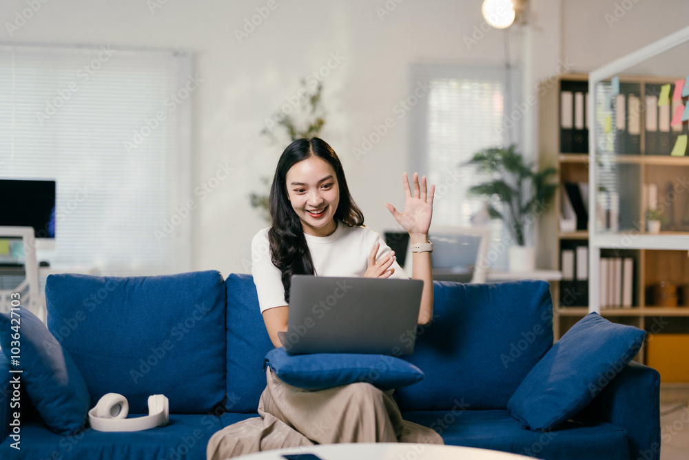 Young asian woman happily waves during a work video call from her cozy living room home office, showcasing the ease of remote work in today's digital world Young asian woman happily waves during a work video call from her cozy living room home office, showcasing the ease of remote work in today's digital world