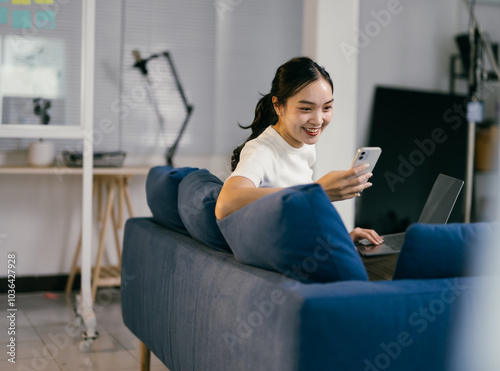 Young woman is working from home, sitting on a comfortable blue sofa and using her laptop and smartphone. She is smiling and seems to be enjoying her work