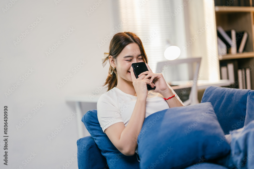 Young woman is spending leisure time in her living room, comfortably seated on a sofa while engaging with her smartphone. She is enjoying her online activity Young woman is spending leisure time in her living room, comfortably seated on a sofa while engaging with her smartphone. She is enjoying her online activity