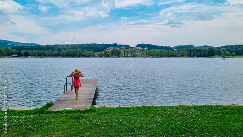 Tourist woman in dress walking on wooden pier extends into the calm waters of the lake Stubenbergsee surrounded by rolling hills of East Styrian Hill Country in Styria, Austria. Forest on foothills photo