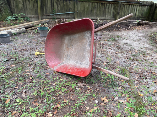 Overturned red wheelbarrow on dirt ground, scattered leaves and tools, backyard garden scene, rustic vibe. 