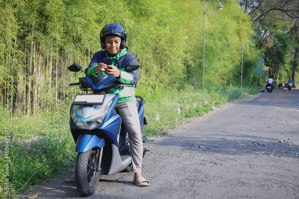 Obraz premium Online taxi rider wearing green jacket and helmet, riding motorcycle, using a mobile phone while on the way to pick up customer. Transportation application concept.