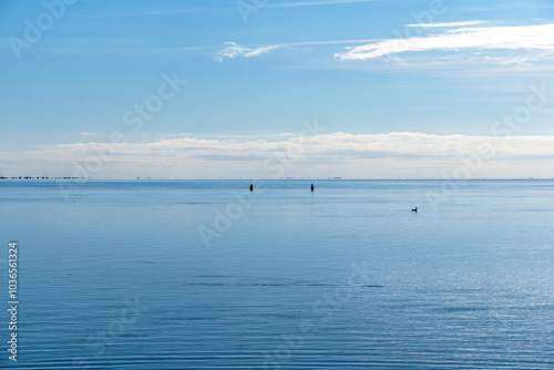 Calm Mediterranean Sea in the Ebro Delta with two backlit fishermen and a duck under a clear blue sky with Trabucador beach on the horizon.