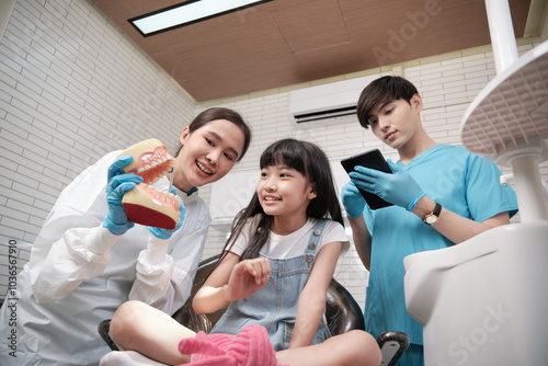 Asian female pediatric dentist and assistant demonstrate toothbrush to little girl with teeth model in dental clinic, well-being hygiene, and professional orthodontic healthcare work in kid hospital.