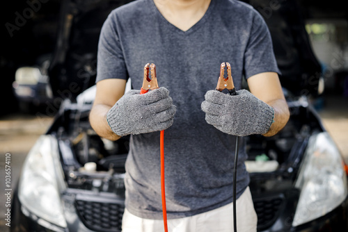 A man stands holding a jump starter cable to charge a dead car battery. Driver standing with jump start cables against a background of a dead car battery.