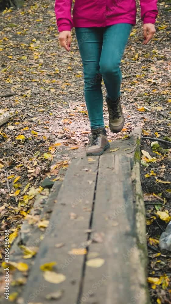 Female legs in jeans and trekking boots walking on a small wooden footbridge in an autumn forest, low-angle front view, slow motion Female legs in jeans and trekking boots walking on a small wooden footbridge in an autumn forest, low-angle front view, slow motion