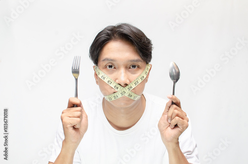 a diet concept illustrated by young asian man with unhappy expression covering his mouth with a measuring tape. eating disorder concept. extreme diet concept. man holding spoon and fork hungrily.