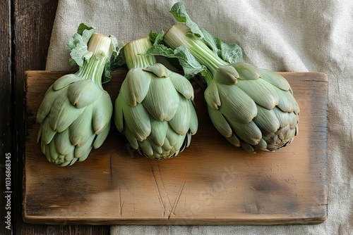 Three fresh artichokes placed on a rustic wooden board, with textured fabric in the background, creating an earthy and organic kitchen scene photo