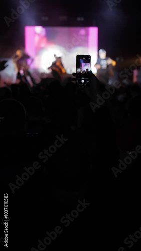 A concert scene featuring a band on stage, with an audience standing, dancing, and waving their hands in the background. Silhouettes of people create an energetic atmosphere.